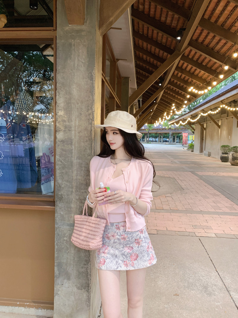 Woman wearing pink cardigan, floral skirt, and beige hat, holding a drink and pink woven bag.