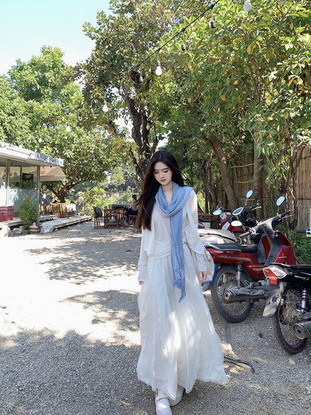 Woman wearing a white flowing dress with a light blue scarf, standing outdoors near trees and scooters.