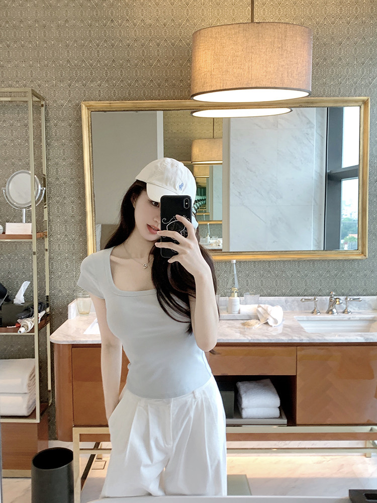 Woman taking mirror selfie wearing light gray top and white pants with white cap