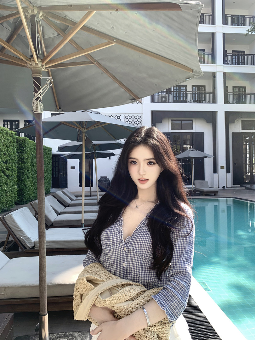 Woman wearing a checkered shirt by a poolside with lounge chairs and umbrellas