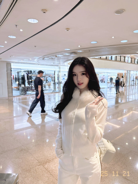 Woman wearing a white knit cardigan with a fur collar and matching white pants in a shopping mall.