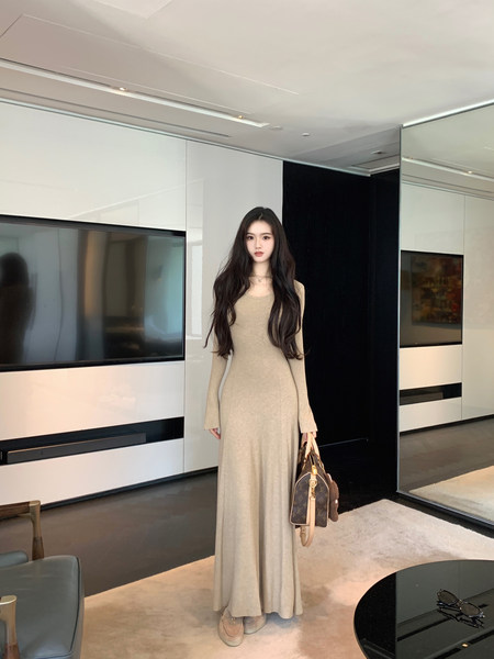 Woman wearing a beige long-sleeve maxi dress, holding a designer handbag in a modern living room.