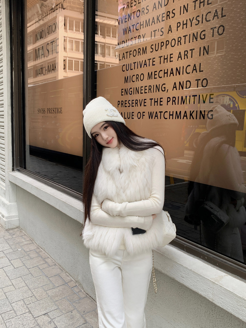 Woman wearing white fur vest, white knit hat, and white pants standing outside a store window.