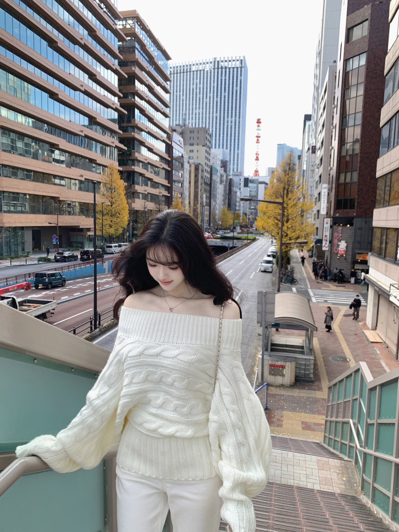 Woman wearing off-shoulder white cable knit sweater and white pants on city balcony
