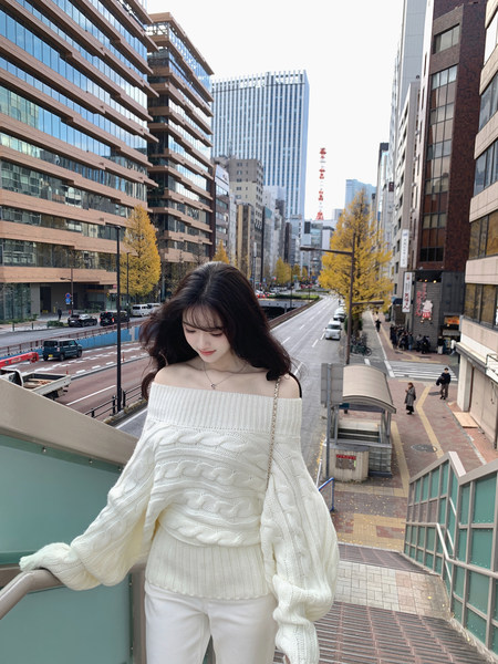 Woman wearing off-shoulder white cable knit sweater and white pants on city balcony