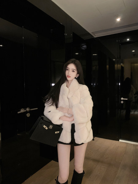 Woman wearing a white faux fur coat with black shorts and boots, holding a black handbag.