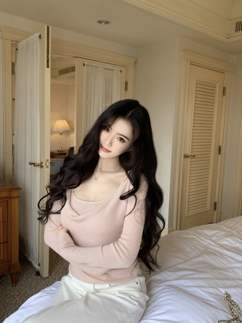Woman wearing a pink ribbed sweater and white pants, sitting on a bed in a hotel room.