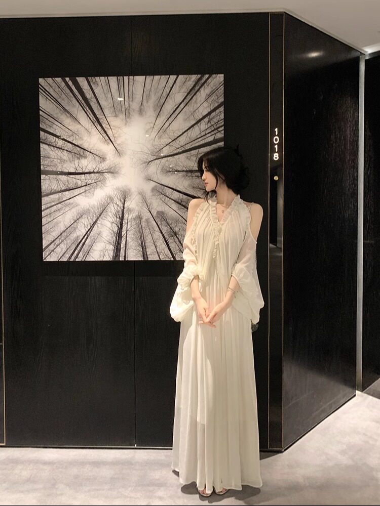 Woman wearing a white long dress with sheer sleeves, standing indoors against a dark wall with artwork.