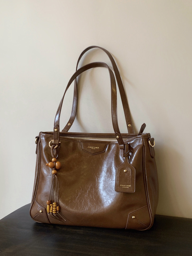Brown leather tote handbag with long straps and decorative tassel charm on a plain background