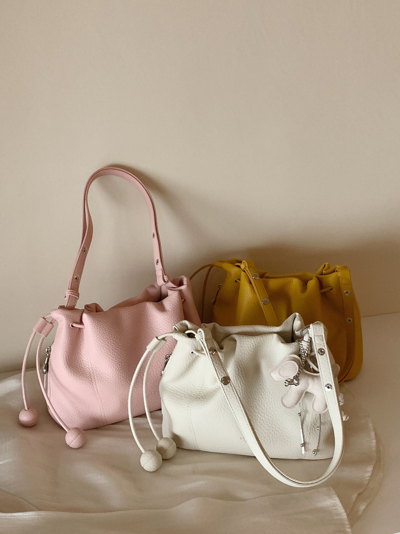Three leather bucket handbags in pink, white, and yellow displayed on a neutral surface