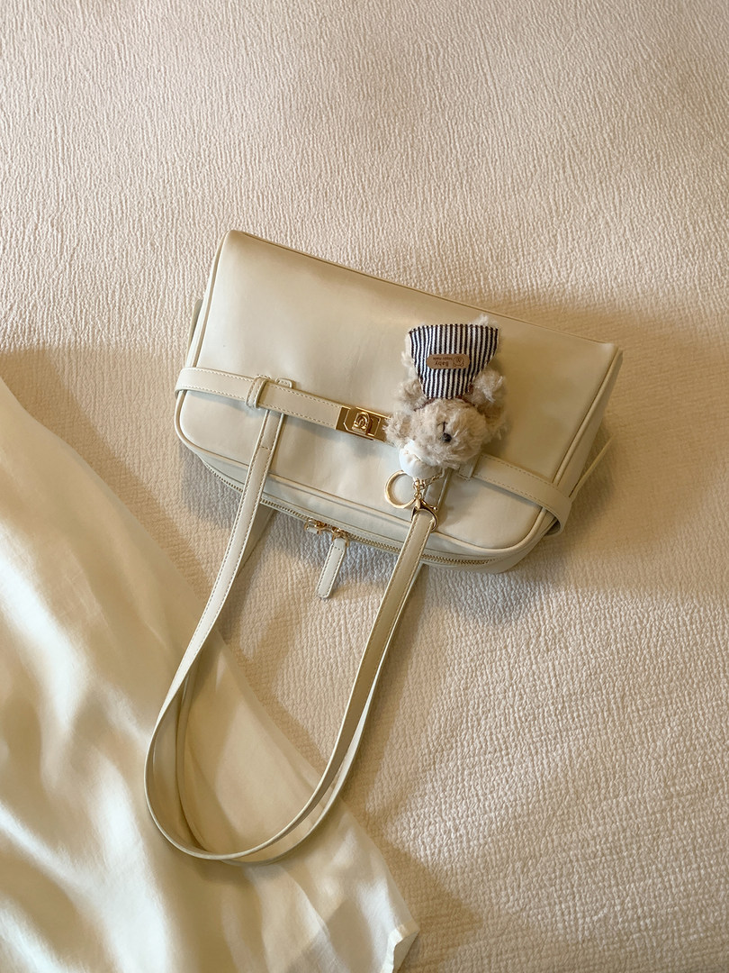 Cream leather shoulder bag with a teddy bear charm on a textured fabric background