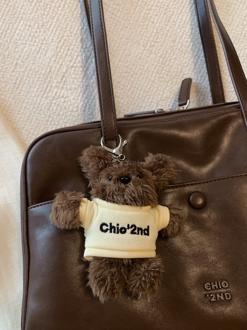 Brown leather handbag with teddy bear charm on a white background