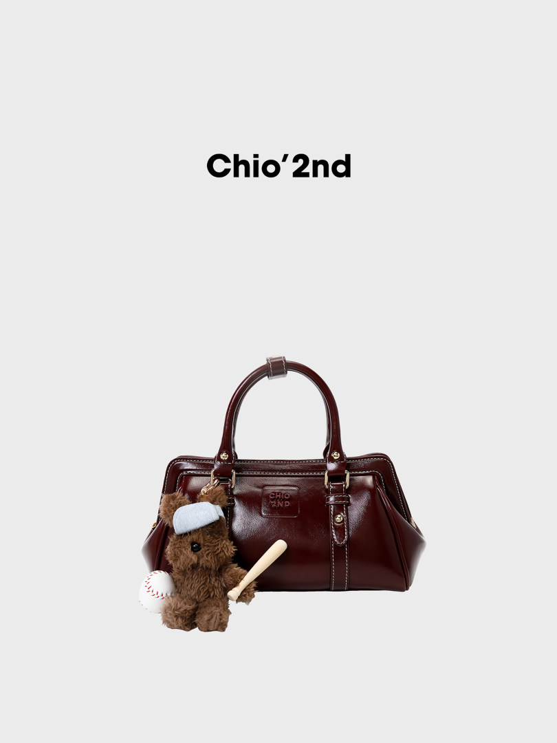 Brown leather handbag with teddy bear charm and baseball accessory on a white background
