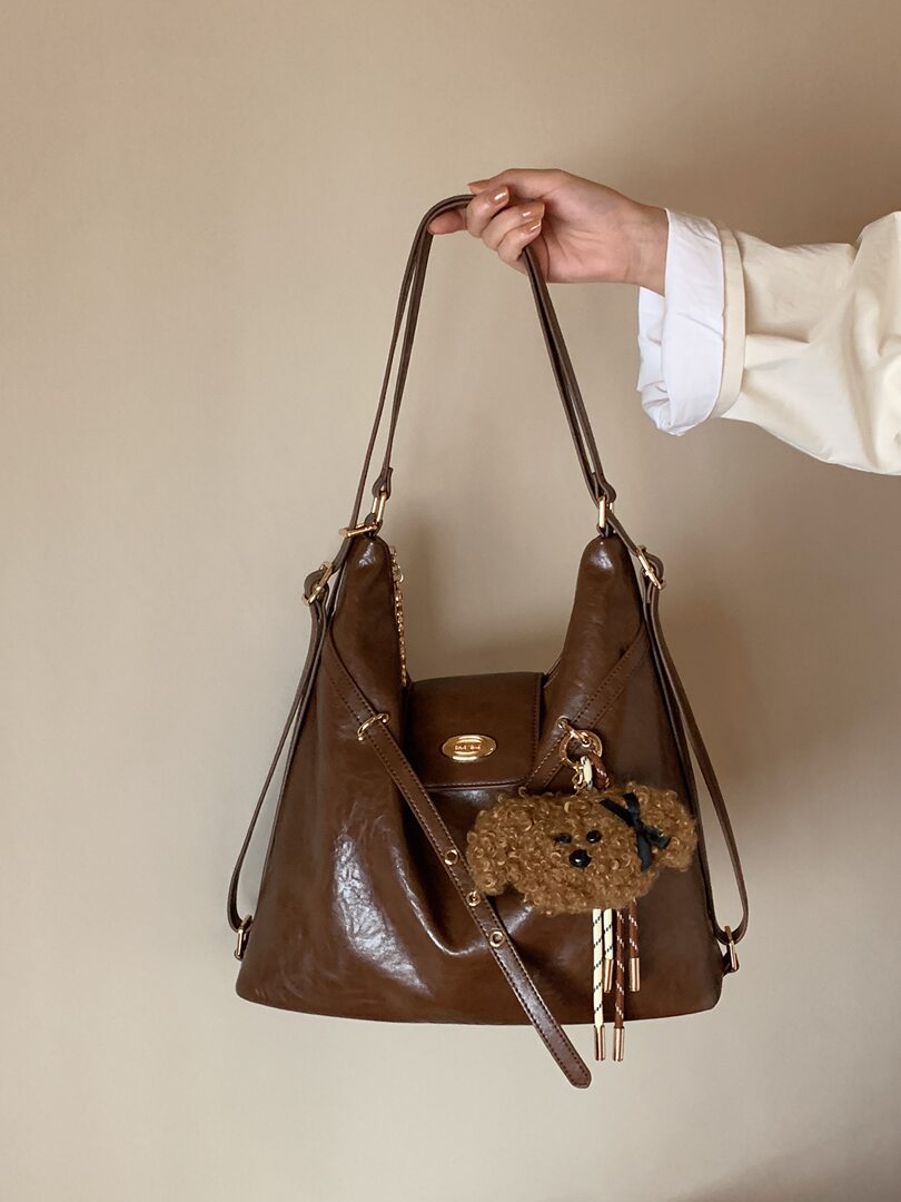 Brown leather shoulder handbag with a teddy bear charm being held by hand