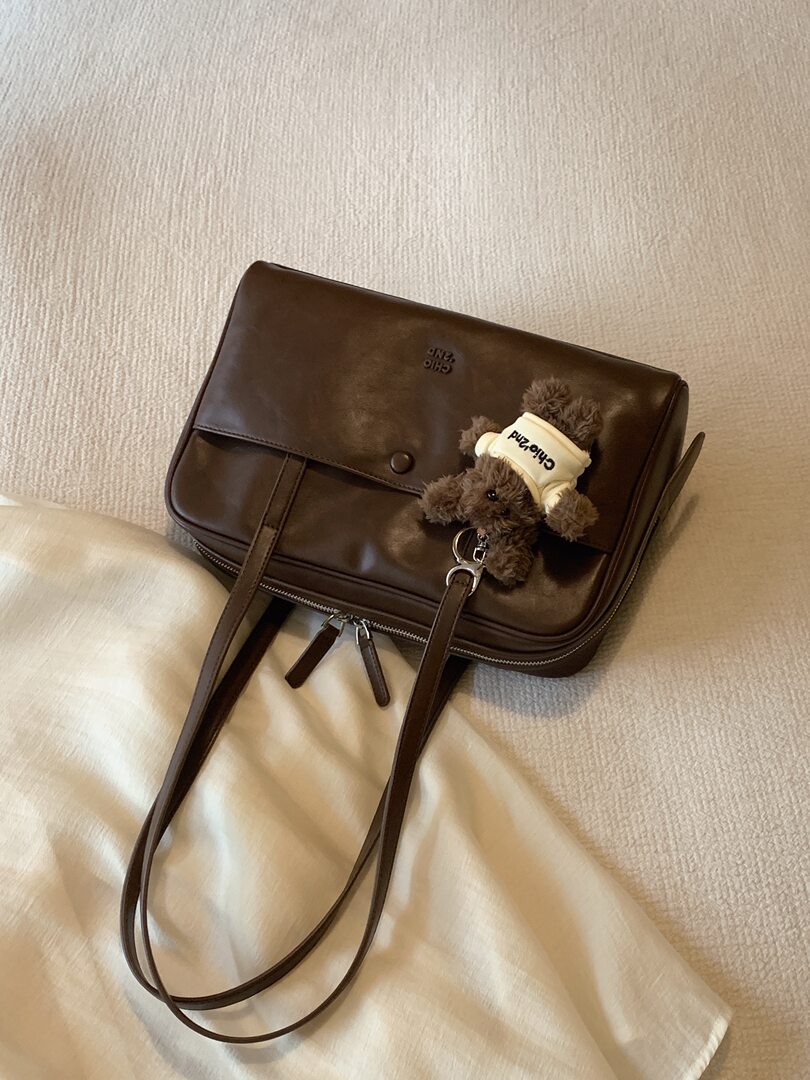 Brown leather shoulder handbag with a teddy bear charm on a bed