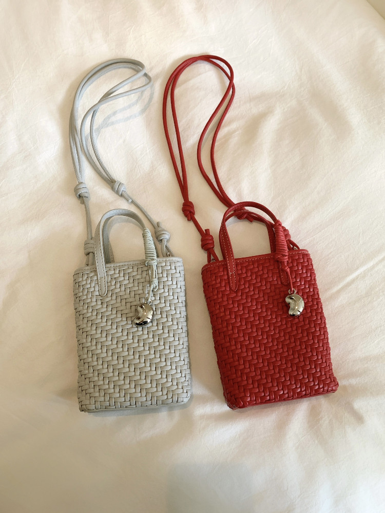 Two small woven mini handbags with long straps, one light gray and one red, displayed on a fabric background