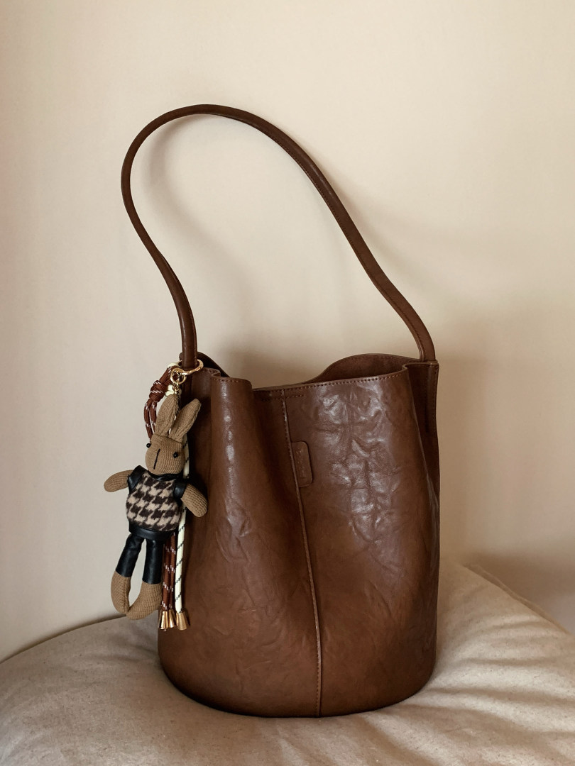 Brown leather bucket handbag with a long shoulder strap and decorative rabbit charm