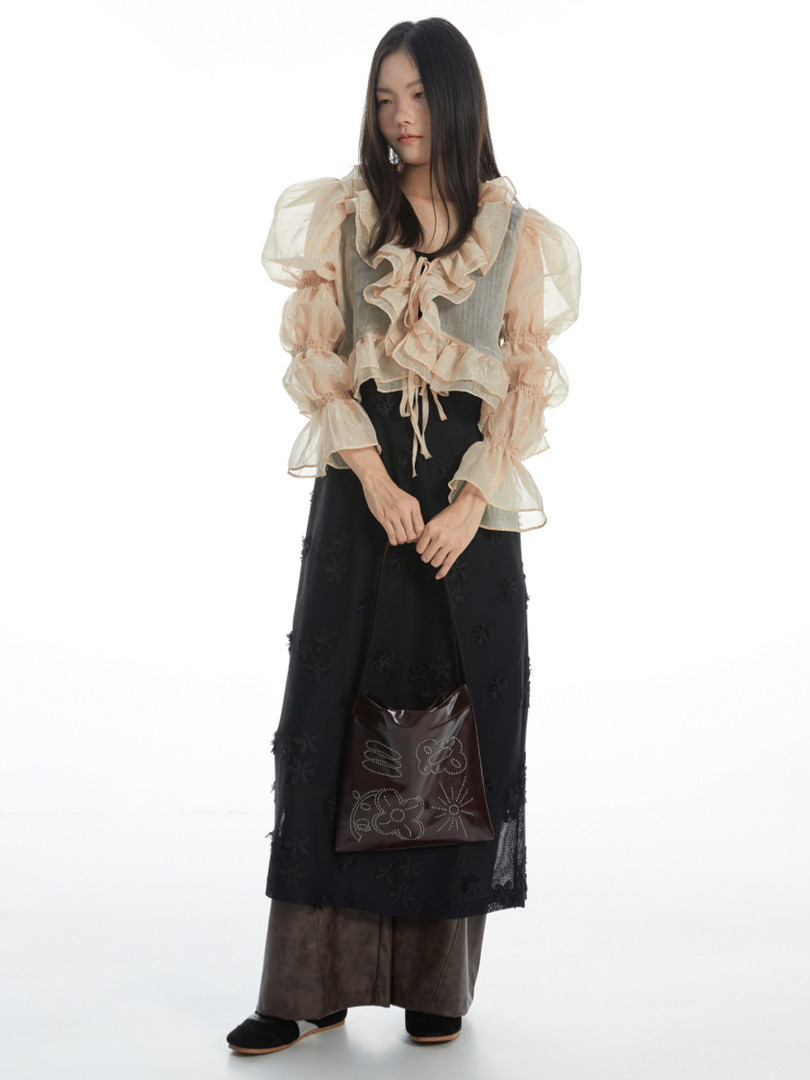 Model wearing a sheer ruffled blouse with a long black skirt and holding a brown handbag