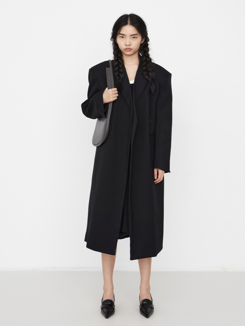 Model wearing a long black oversized coat with a shoulder bag and black flats