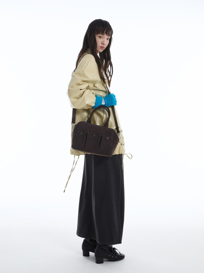 Model wearing a dark shoulder bag with a beige jacket and black skirt in a studio setting