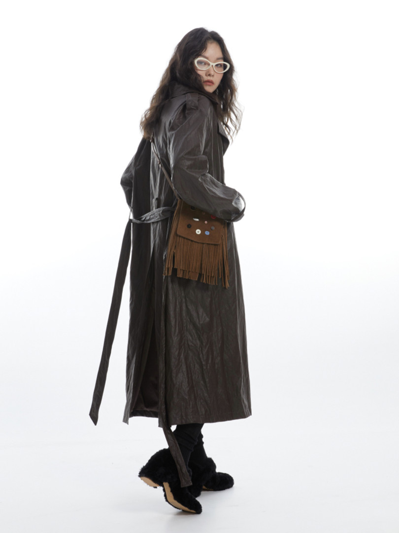 Model wearing a long dark trench coat with a fringe crossbody bag and black boots on a white background