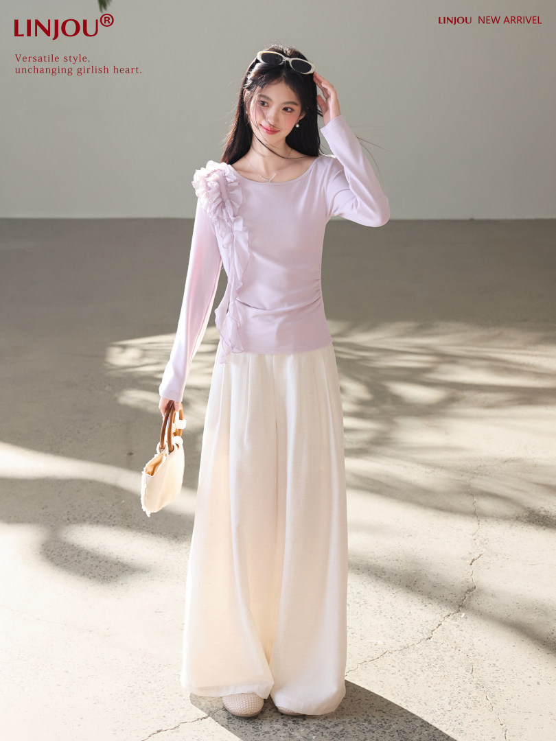 Model wearing a pastel pink long-sleeve top with a cream wide-leg skirt or pants, holding a small handbag