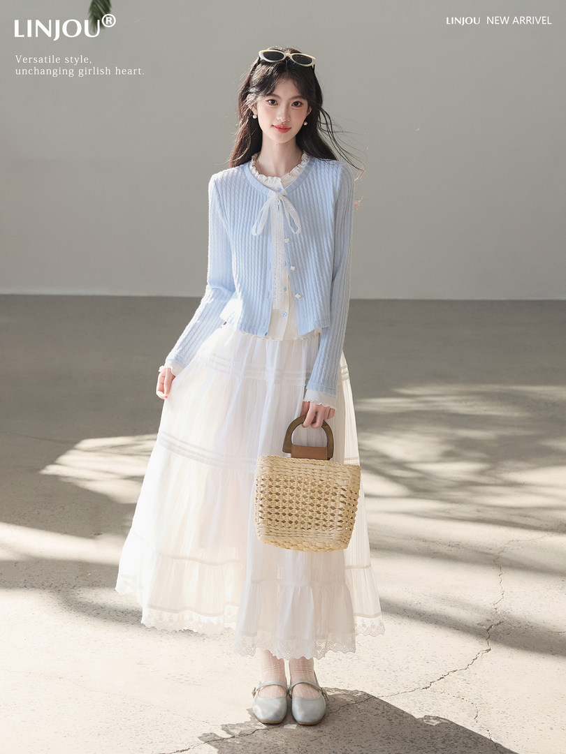 Model wearing a light blue cardigan over a white long skirt with a woven handbag