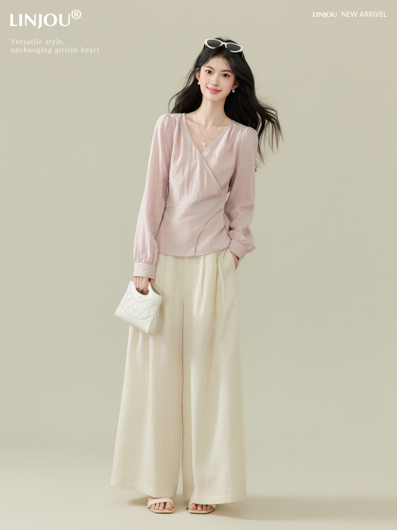 Model wearing a light pink wrap blouse with cream wide-leg pants and a small white handbag