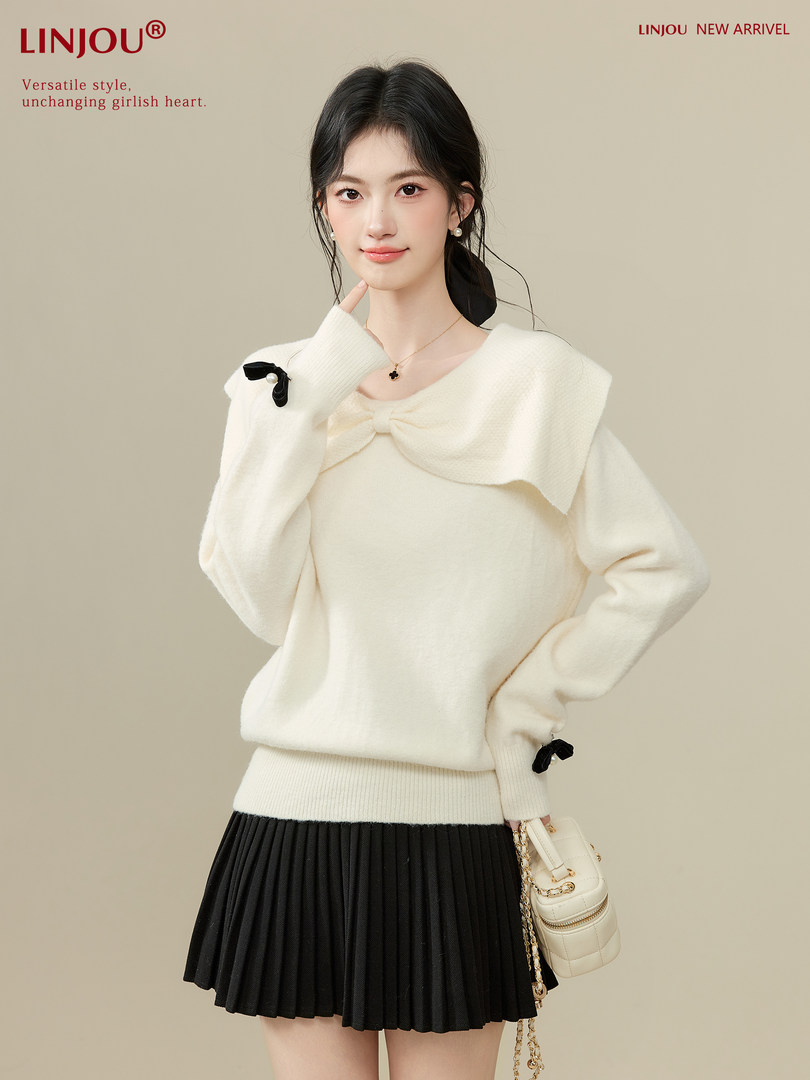 Model wearing a cream knit sweater with a black pleated skirt and small handbag