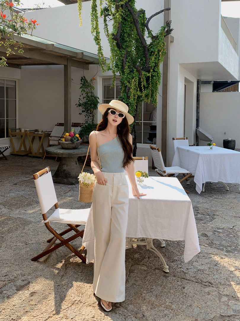 Woman wearing a light blue one-shoulder top with cream wide-leg pants and a straw hat