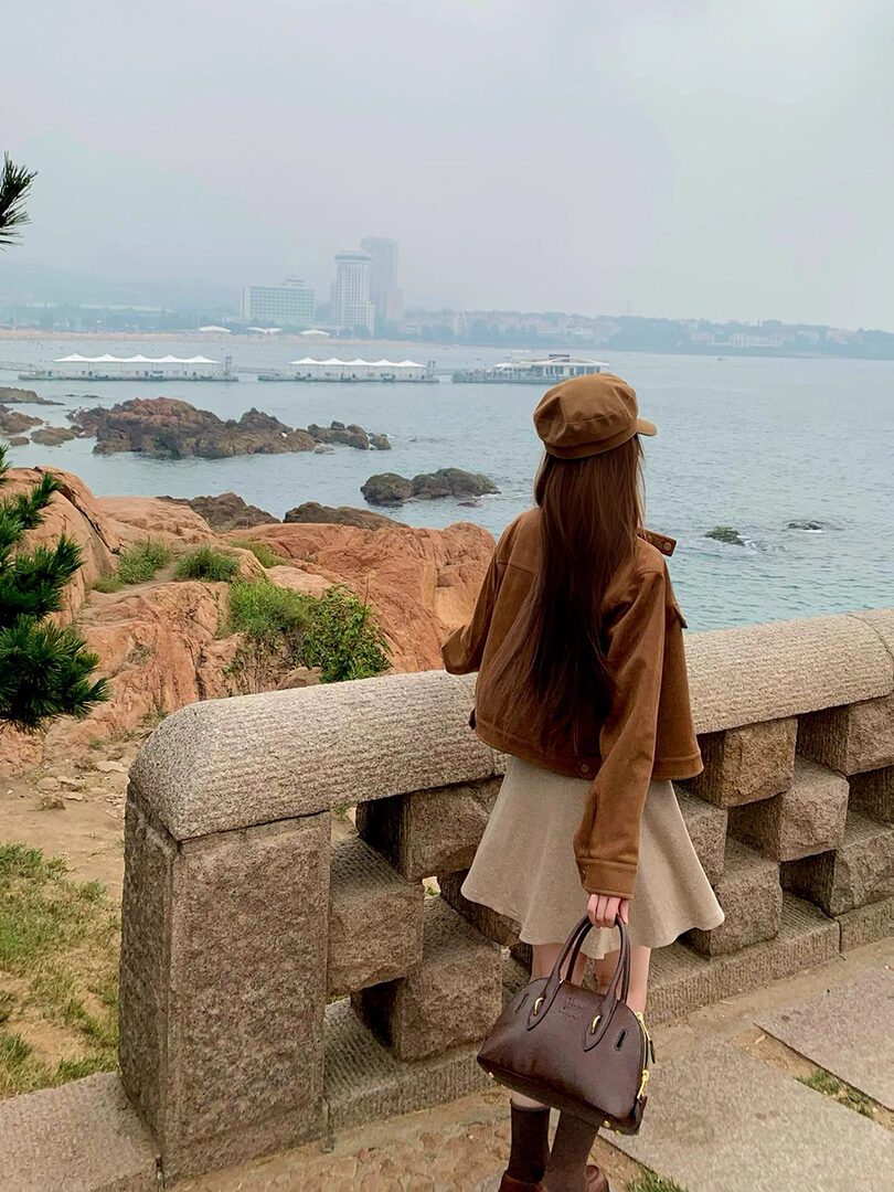 Woman wearing a brown jacket and beige skirt by the seaside while holding a handbag