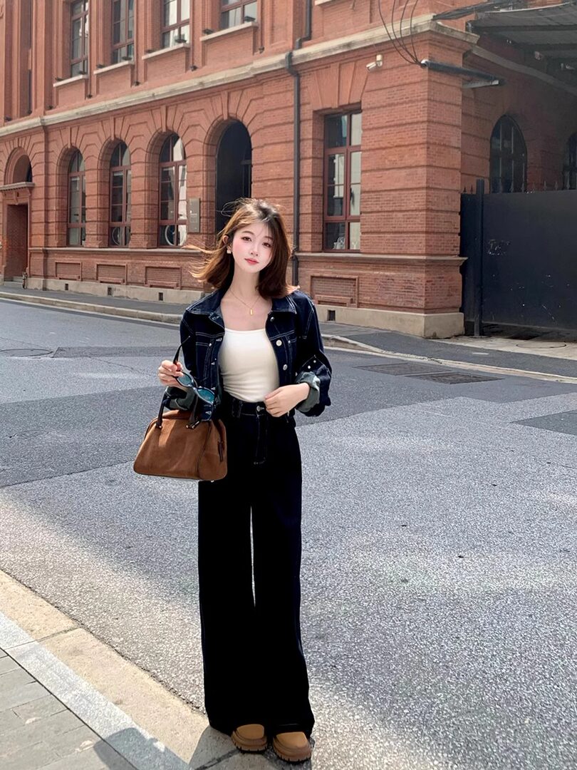 Woman wearing a dark denim jacket with black wide-leg pants and a brown handbag on a city street