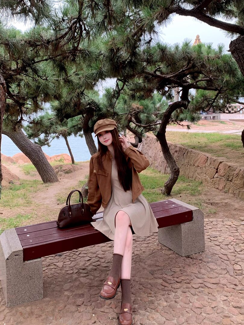 Woman wearing a brown jacket, beige dress, loafers, and cap outdoors on a bench