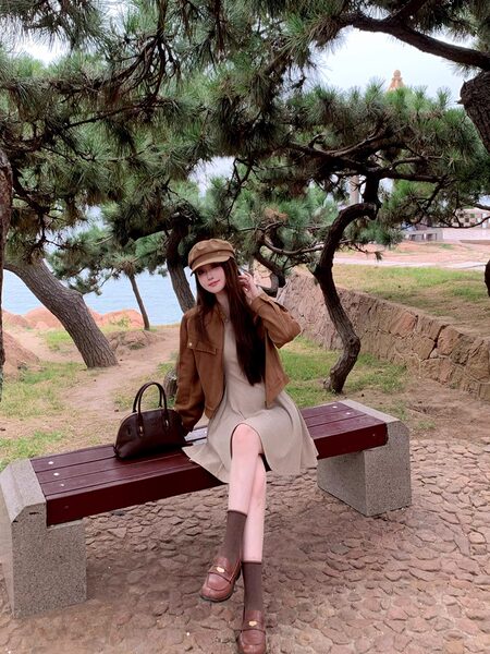 Woman wearing a brown jacket, beige dress, loafers, and cap outdoors on a bench