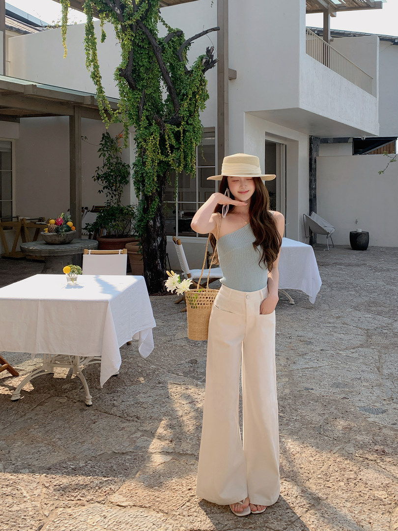 Woman modeling a light blue one-shoulder top with white wide-leg pants, straw hat, and woven bag