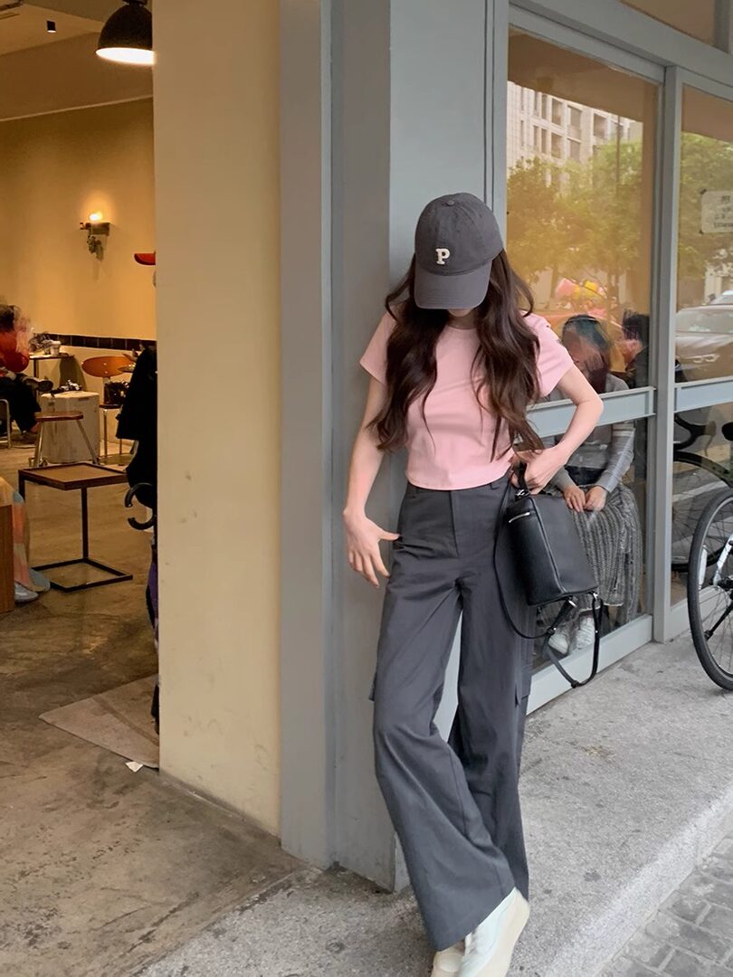 Woman wearing a pink T-shirt, gray wide-leg pants, and a black cap with a handbag