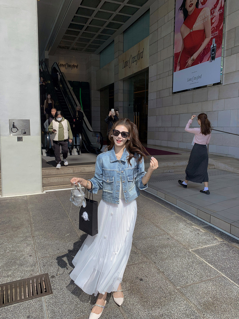 Woman wearing a light blue denim jacket with a white pleated maxi skirt and flats outdoors