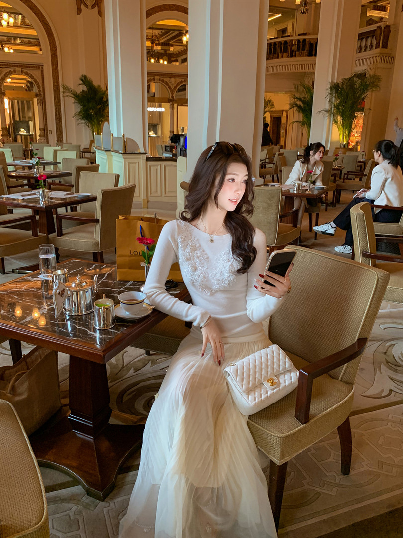 Woman wearing a white embroidered top and sheer pleated skirt with a quilted white handbag in a luxury restaurant.