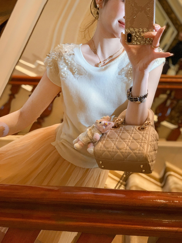 Woman wearing a white embellished top and holding a quilted beige handbag in a mirror selfie