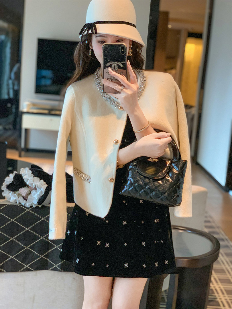 Mirror selfie of a woman wearing a cream cape jacket over a black embellished dress and holding a black quilted handbag