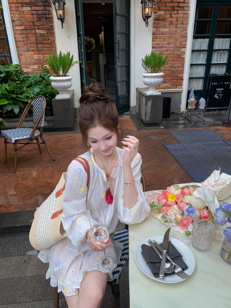 Woman wearing a white floral long-sleeve dress with a straw tote bag at an outdoor cafe