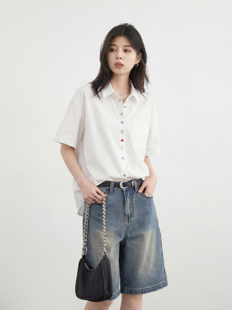 Model wearing a white short-sleeve button-up shirt with denim shorts and a black shoulder bag