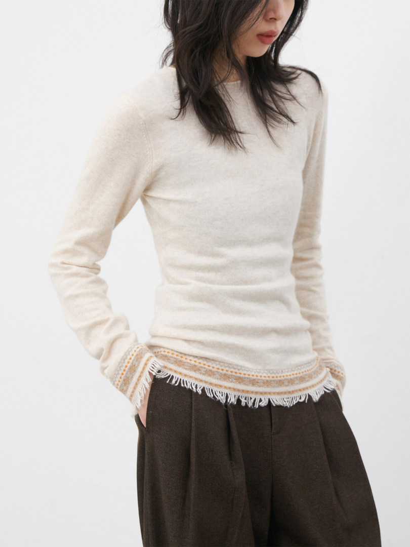 Model wearing a cream long-sleeve knit top with fringe hem and dark pants