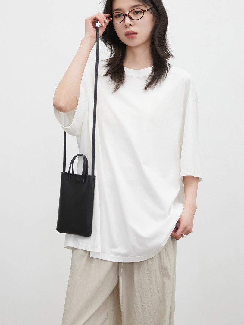 Model wearing an oversized white short-sleeve top and holding a black crossbody mini bag