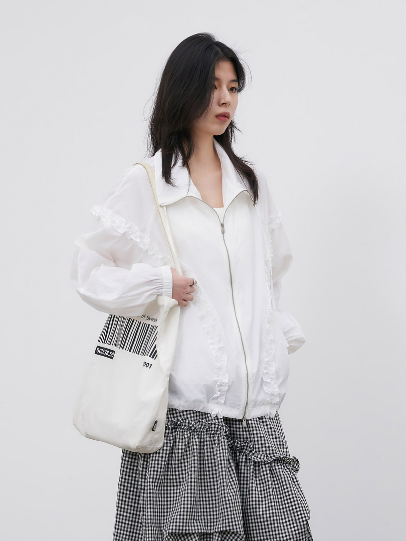 Model wearing a white zip-up jacket with ruffled details and a black-and-white checkered skirt, carrying a tote bag