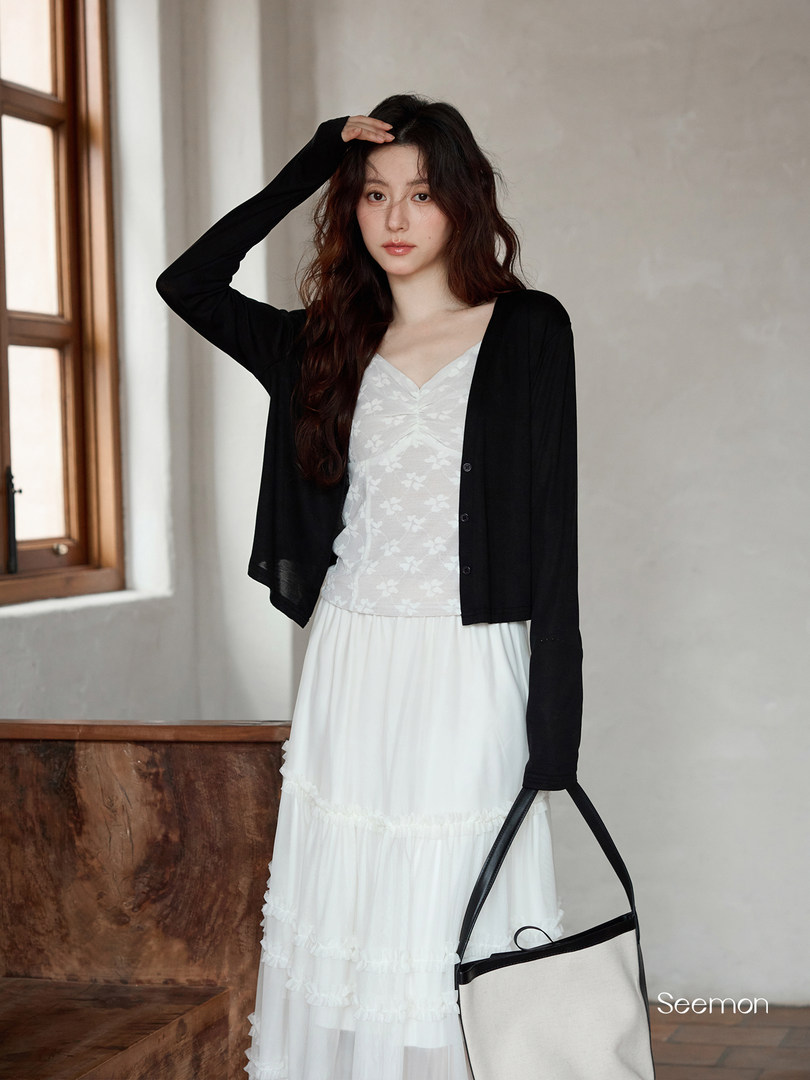 Model wearing a black cardigan over a white tiered dress with a tote bag