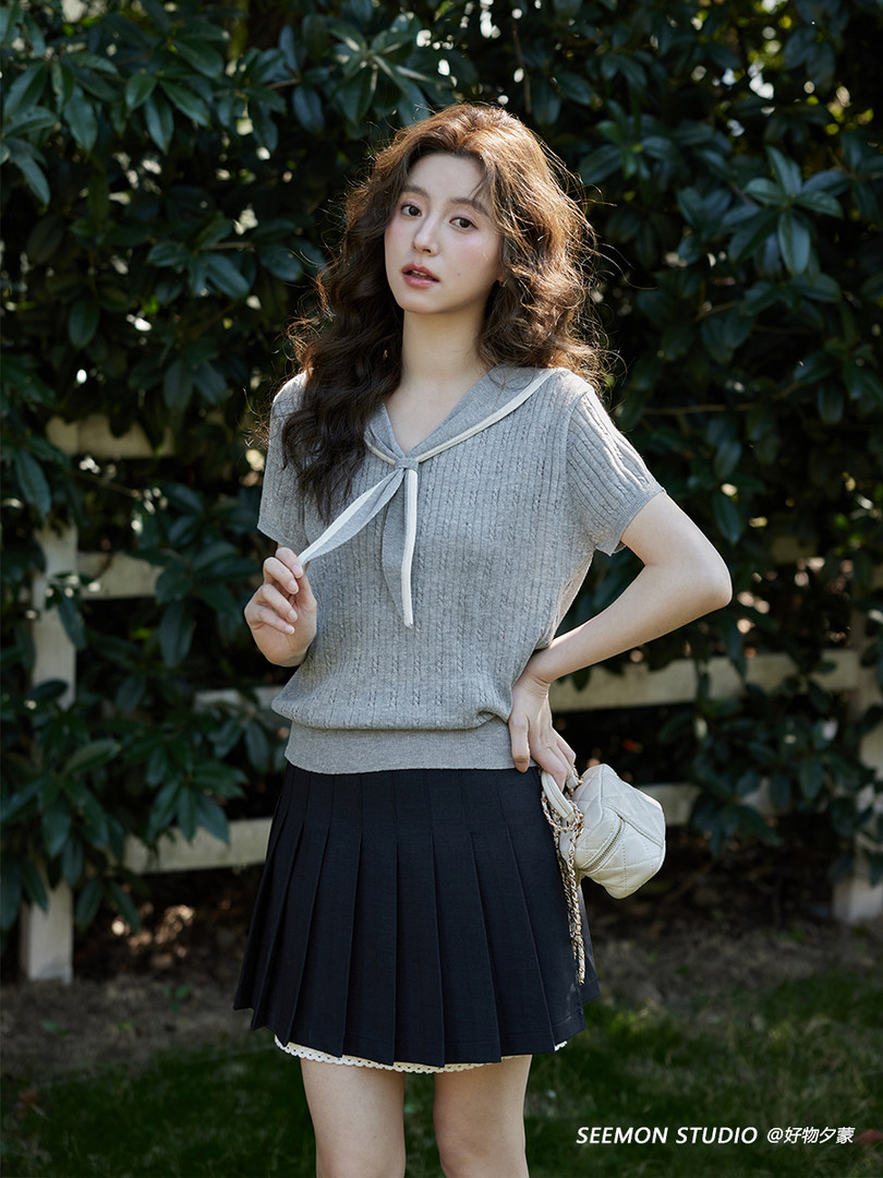 Model wearing a gray short-sleeve knit top with a sailor-style tie and black pleated skirt outdoors