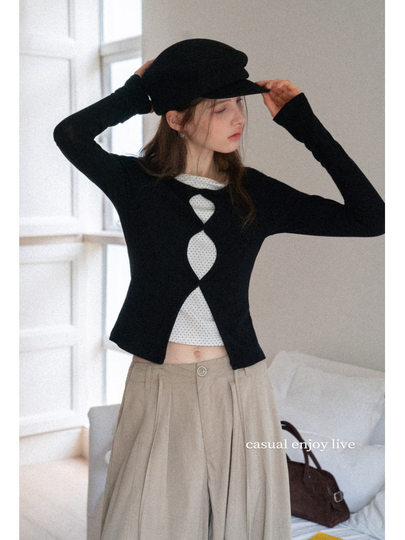 Model wearing a black long-sleeve top with white cutout detail, beige wide-leg pants, and a black cap