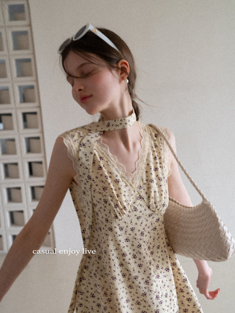 Model wearing a cream floral sleeveless dress with lace trim and a woven shoulder bag