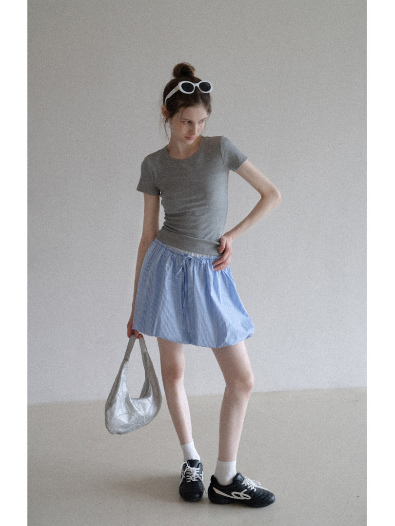 Model wearing a gray fitted T-shirt, light blue skirt, sneakers, and holding a silver bag